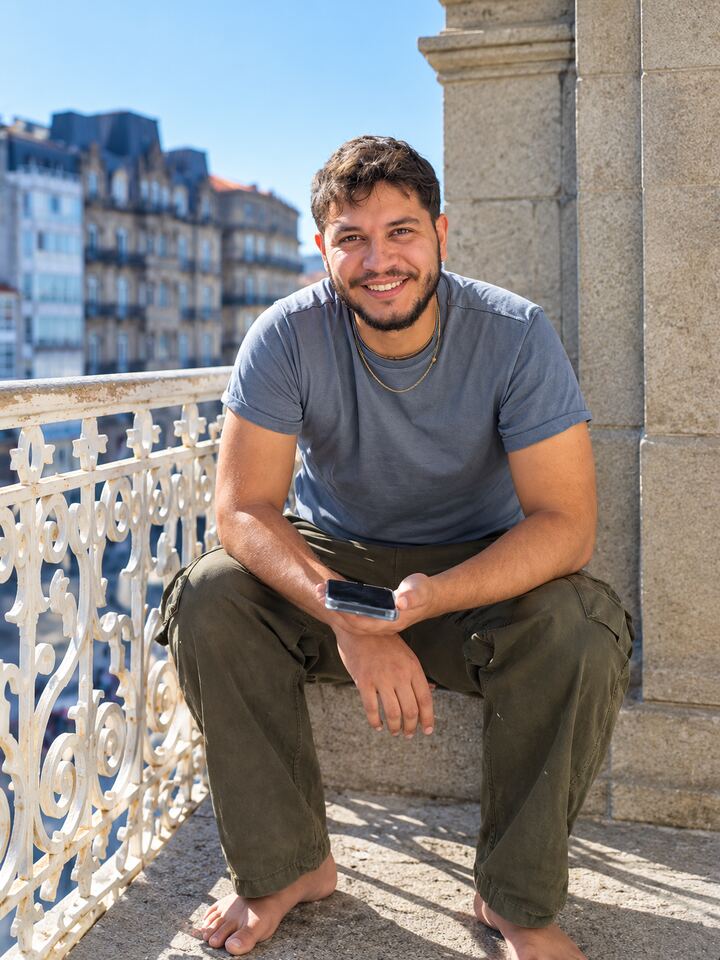 Felipe, smiling, sits on a sunlit stone balcony in A Coruña with a phone in his hands. An ornate white iron railing and tall old buildings are behind him.